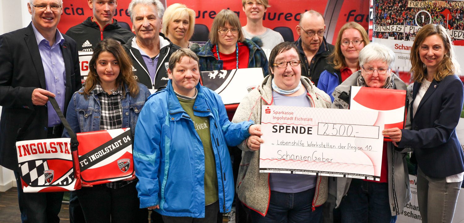 Eine Gruppe von Menschen hält einen Scheck über 2500 Euro von der Sparkasse Ingolstadt Eichstätt für die Lebenshilfe Werkstätten der Region 10. Die Personen stehen vor einem roten Hintergrund mit dem Logo des FC Ingolstadt 04 und halten Trikot-Taschen mit dem Vereinslogo. Die Initiative heißt "SchanzenGeber".
