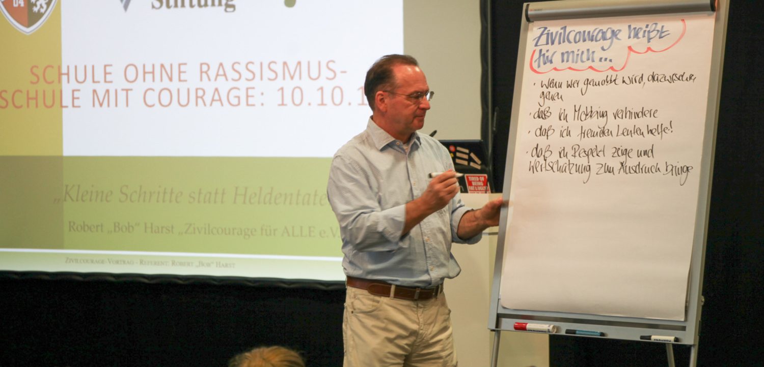 Ein Mann steht vor einer Gruppe von Zuhörern und hält einen Vortrag über Zivilcourage. Er schreibt auf eine Flipchart, auf der bereits der Titel "Zivilcourage heißt für mich..." zu lesen ist. Im Hintergrund ist eine Präsentation mit dem Logo des FC Ingolstadt 04, der Hanns Seidel Stiftung und dem Titel "Schule ohne Rassismus - Schule mit Courage: 10.10.10" zu sehen. Der Mann trägt ein blaues Hemd und beigefarbene Hosen.