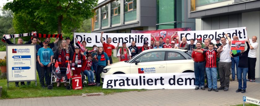 Eine Gruppe von Menschen steht vor einem Gebäude und hält ein großes Transparent, auf dem "Die Lebenshilfe gratuliert dem FC Ingolstadt!" steht. Die Menschen tragen verschiedene T-Shirts und Schals in den Farben des FC Ingolstadt und posieren mit einem Auto, das mit einem Lebenshilfe-Logo versehen ist. Im Hintergrund ist ein Schild mit der Aufschrift "Lebenshilfe Werkstätten" zu sehen.