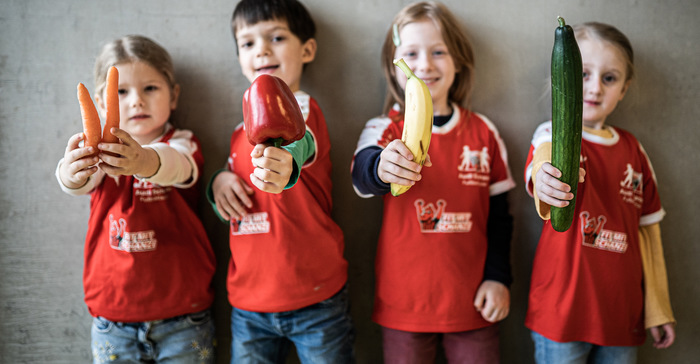Vier Kinder in roten T-Shirts mit einem Logo halten verschiedene Gemüsesorten und Obst in die Kamera. Ein Kind hält eine Karotte, ein anderes eine Paprika, das dritte Kind hält eine Banane und das vierte Kind einen Gurke.