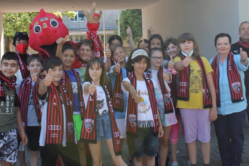 Eine Gruppe von Kindern und Erwachsenen steht vor einem Gebäude und hält Schals mit rot-schwarzen Streifen und dem Logo des 1. FC Köln in die Kamera. Einige der Kinder tragen T-Shirts mit Aufdrucken und Accessoires. Ein Kind hält ein rotes Maskottchen mit großen Augen und Ohren hoch. Alle lächeln und zeigen Daumen hoch.