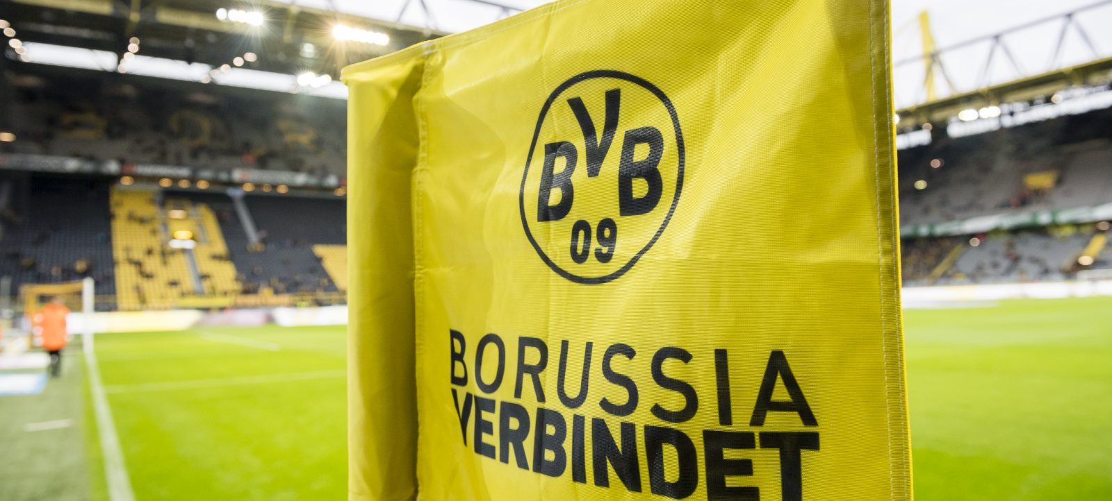 Gelbes Eckfahnen-Tuch mit dem Logo und dem Schriftzug 'BVB 09 Borussia verbindet' im Stadion von Borussia Dortmund. Im Hintergrund ist das leere Stadion mit grünem Rasen und Sitzplätzen zu sehen.