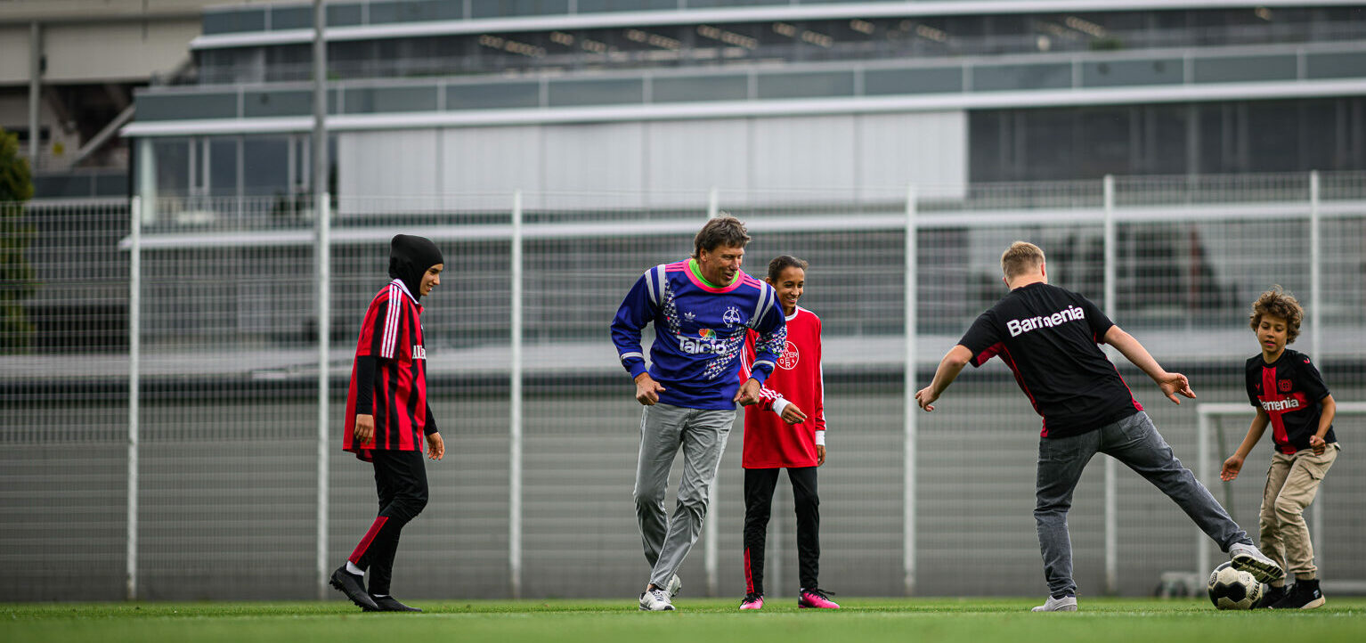 Eine Gruppe von Kindern und einem Erwachsenen auf einem Fußballfeld. Die Kinder tragen verschiedene Trikots und sind in einer lockeren Formation aufgestellt. Ein Kind spielt mit einem Fußball, während die anderen zuschauen. Im Hintergrund ist ein Zaun und ein Gebäude zu sehen.