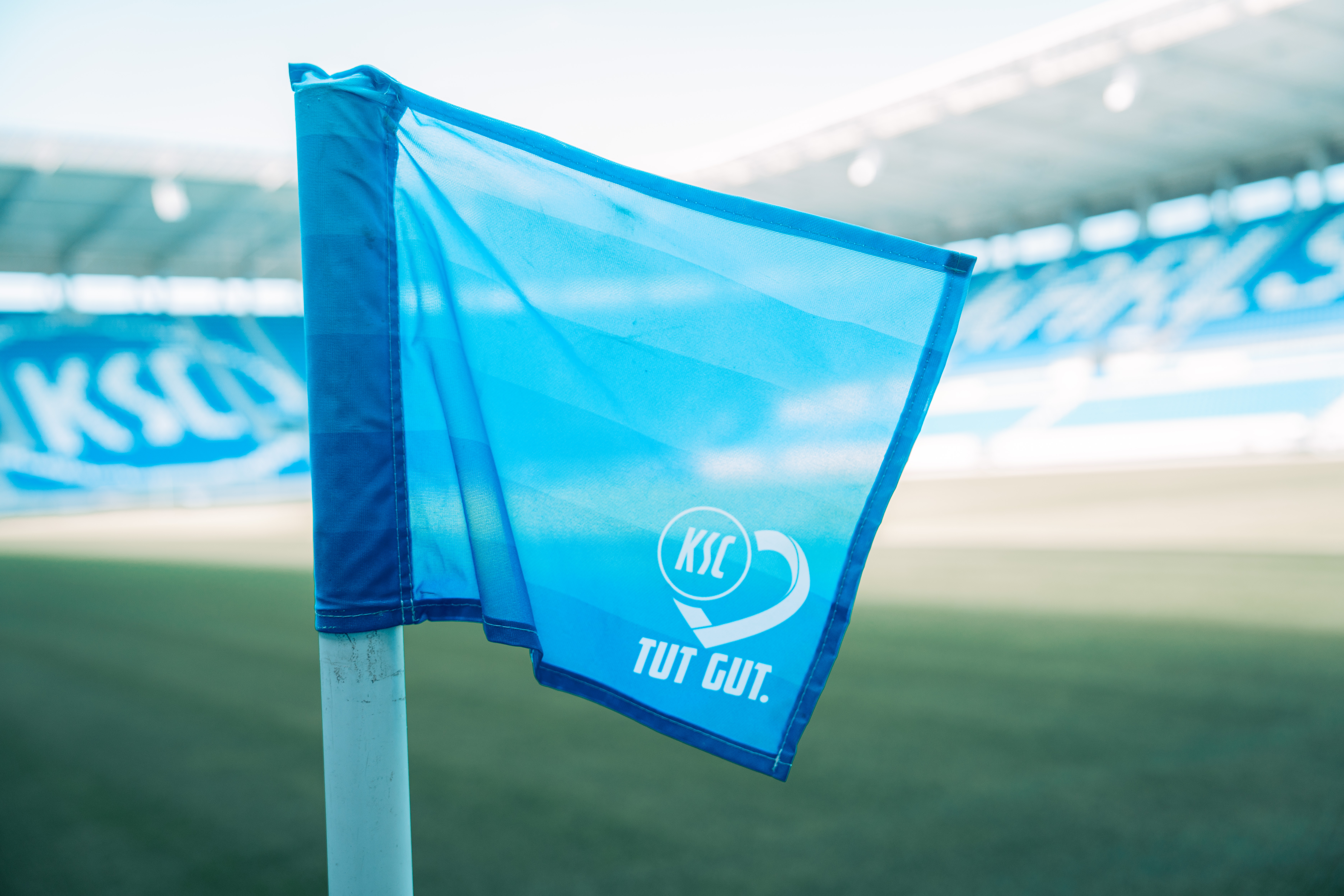 Eine blaue Fahne mit dem Logo des KSC und dem Slogan "TUT GUT." weht auf einem Fußballfeld. Im Hintergrund ist eine Tribüne mit dem Schriftzug "WILD PARK" zu sehen.