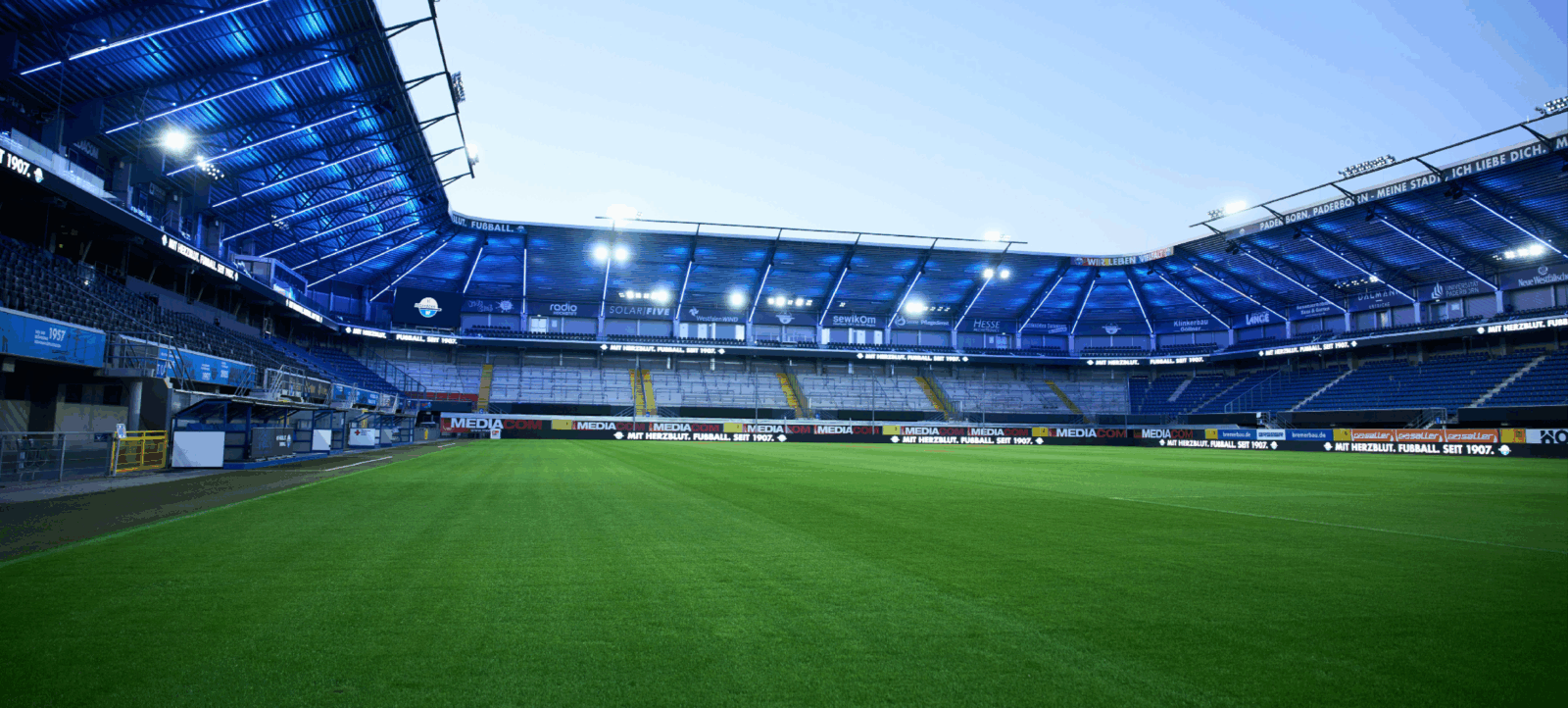 Ein leeres Fußballstadion bei Abendlicht, mit beleuchteten Tribünen und Werbebannern. Der Rasen ist grün und gut gepflegt.