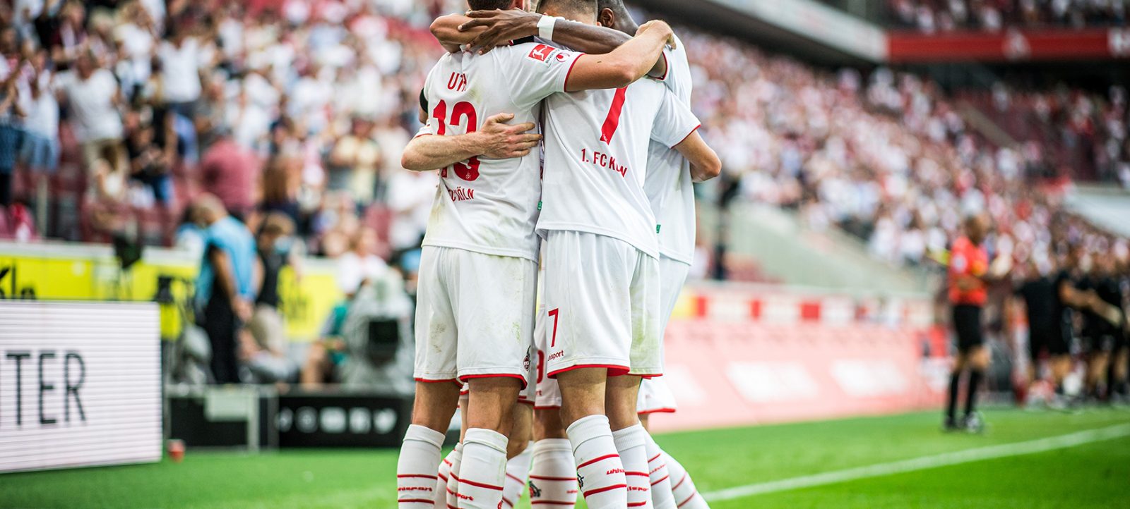 Drei Fußballspieler des 1. FC Köln in weißen Trikots mit roten Akzenten umarmen sich auf dem Spielfeld, um ein Tor zu feiern. Die Spieler stehen vor einem vollen Stadion mit jubelnden Fans. Die Szene spielt im RheinEnergieStadion in Köln während eines Bundesliga-Spiels.