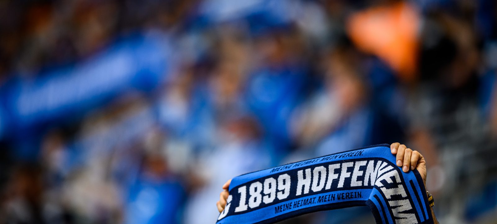 Ein Fan hält ein blaues TSG Hoffenheim-Schal mit dem Text "1899 Hoffenheim - Meine Heimat. Mein Verein." vor einem unscharfen Hintergrund, der vermutlich andere Fans und das Stadion während eines Bundesliga-Spiels zeigt.