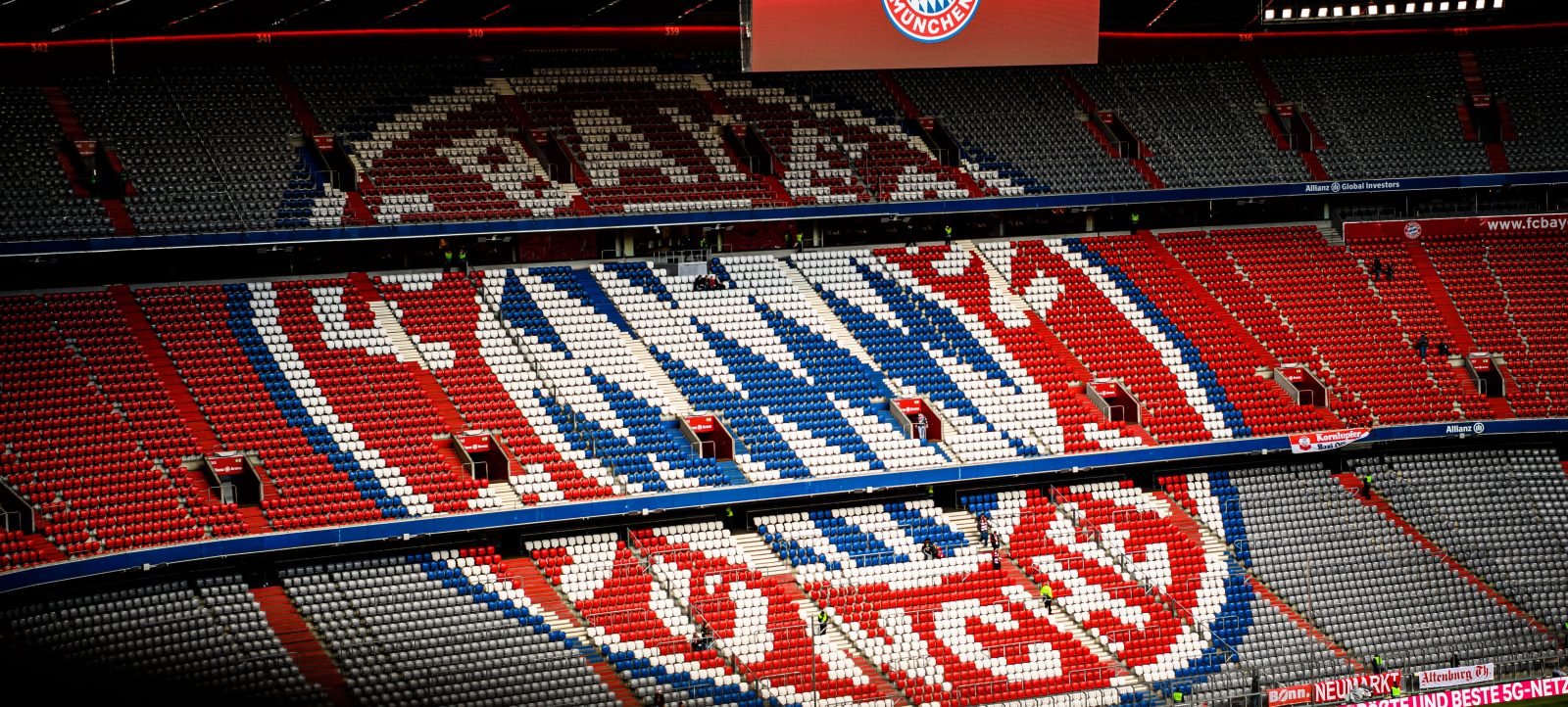 Allianz Arena in München vor einem Bundesliga-Spiel zwischen FC Bayern München und TSG Hoffenheim. Die Tribünen zeigen ein Muster in den Vereinsfarben Rot und Blau mit Weiß. Im Hintergrund ist das Logo des FC Bayern München auf einer großen Anzeigetafel zu sehen.