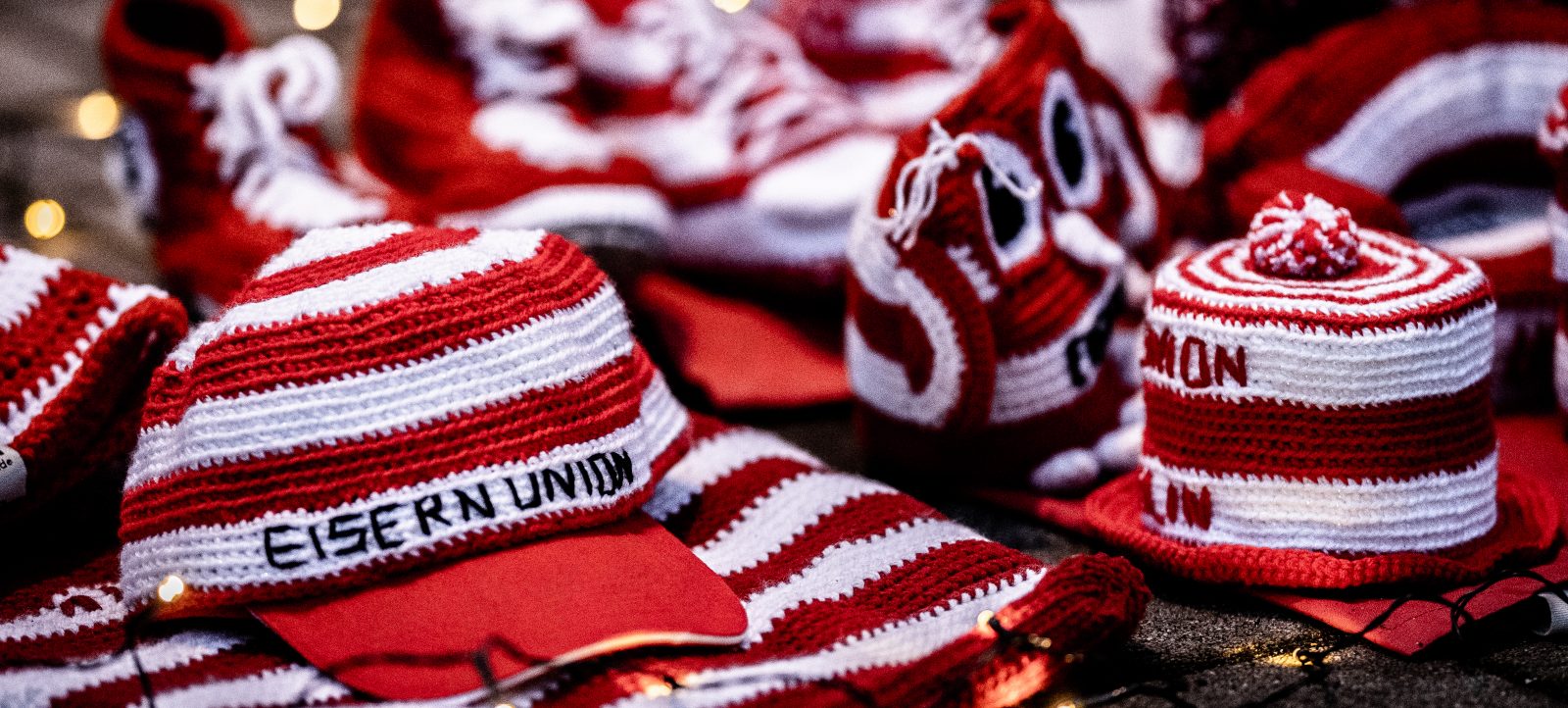 Mehrere rot-weiße gestrickte Mützen mit dem Aufdruck 'Eisern Union', die auf einem Tisch ausgebreitet sind. Die Mützen sind Teil einer Aktion oder eines Verkaufs im Stadion An der Alten Försterei vor einem Bundesliga-Spiel zwischen 1. FC Union Berlin und Hertha BSC.