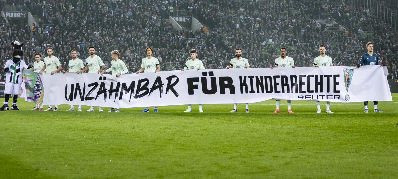 Eine Gruppe von Fußballspielern steht auf dem Platz, sie halten ein Banner hoch: Unzähmbar für Kinderrechte