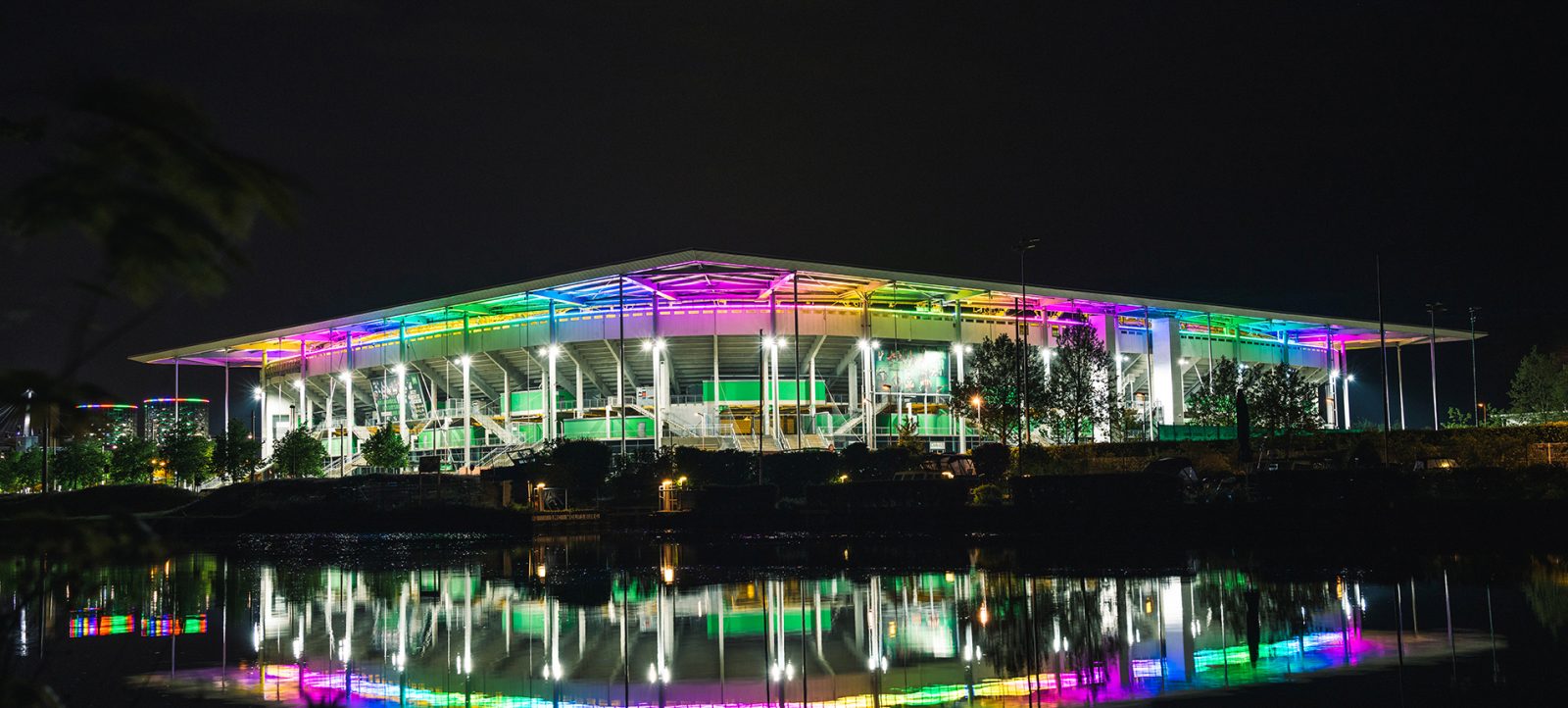 Ein Fußballstadion, die Volkswagen Arena, nachts beleuchtet mit farbenfrohen Lichtern in Regenbogenfarben, die sich im Wasser vor dem Stadion spiegeln.
