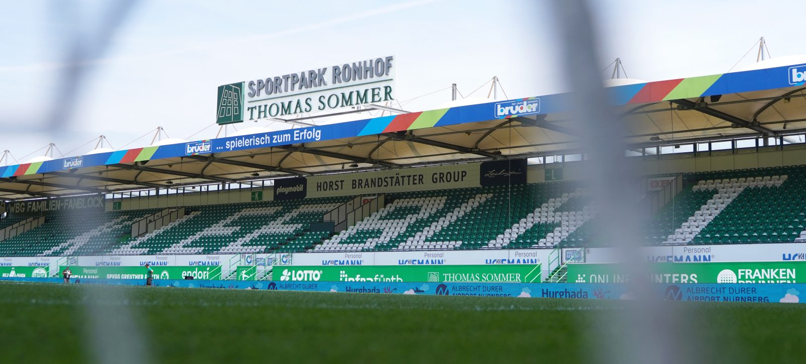 Ein Fußballstadion mit Blick auf die Tribüne und das Spielfeld. Die Tribüne trägt den Namen "Sportpark Ronhof Thomas Sommer" und zeigt verschiedene Werbebanner von Sponsoren wie "bruder", "Horst Brandstätter Group" und "Spielend zum Erfolg". Die Sitzplätze sind grün und weiß gestaltet, und das Bild scheint durch einen leichten Nebel oder Rauch leicht unscharf zu sein.