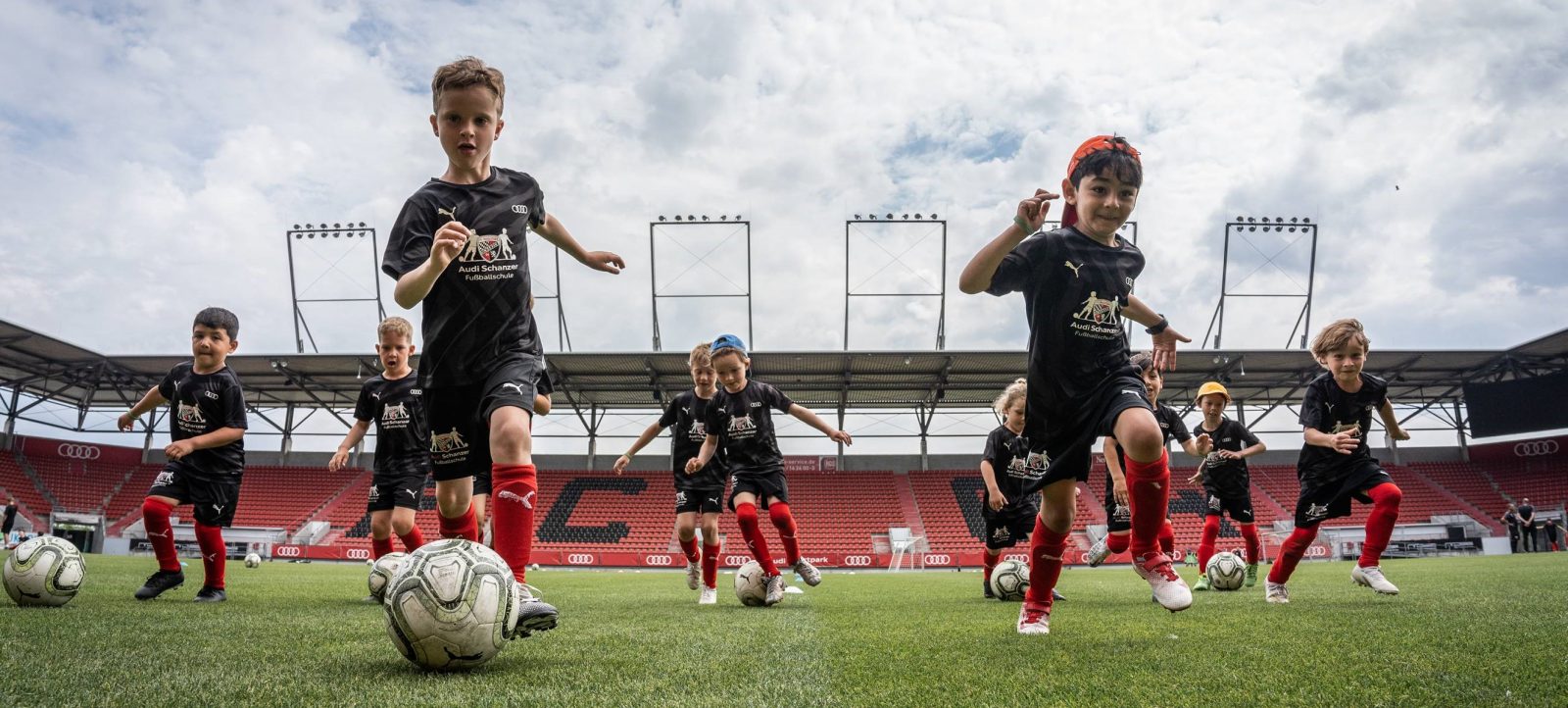 Kinder in einer Fußballschule auf einem Rasenplatz, im Hintergrund ein Stadion mit roten Sitzen und Audi-Werbung.