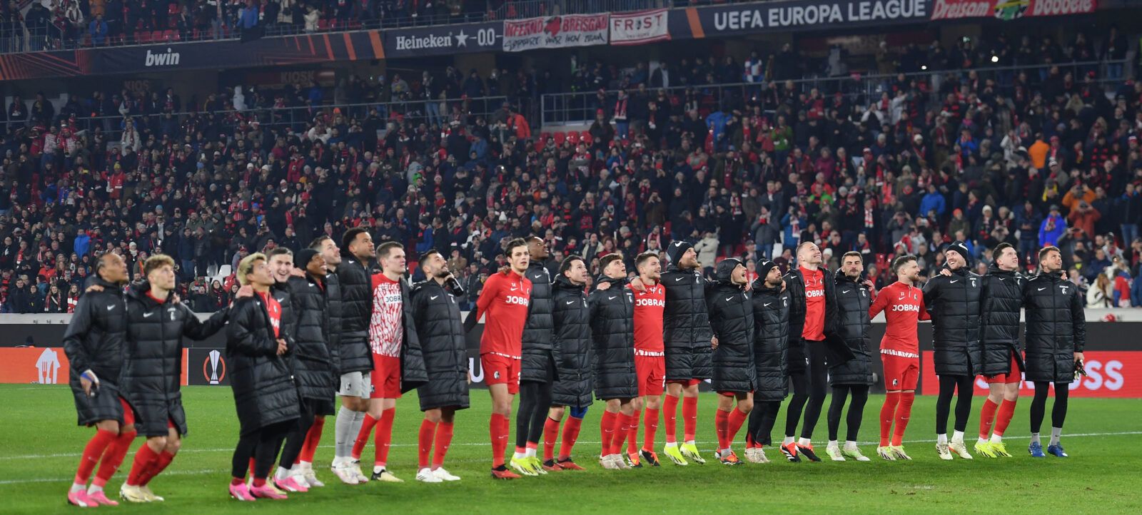 Die Mannschaft des SC Freiburg steht im Europa-Park Stadion und feiert einen Sieg in der UEFA Europa League gegen West Ham. Die Spieler tragen schwarze Jacken und rote Hosen, einige haben die Arme um die Schultern der anderen gelegt. Im Hintergrund sind zahlreiche Zuschauer auf den Tribünen zu sehen.