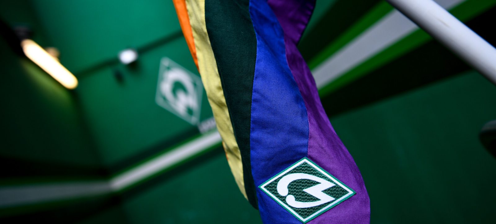 Ein lila und blaues Tuch mit dem Logo des SV Werder Bremen hängt an einem weißen Pfosten auf einem grünen Fußballfeld. Im Hintergrund sind weitere Logos des Vereins zu sehen.