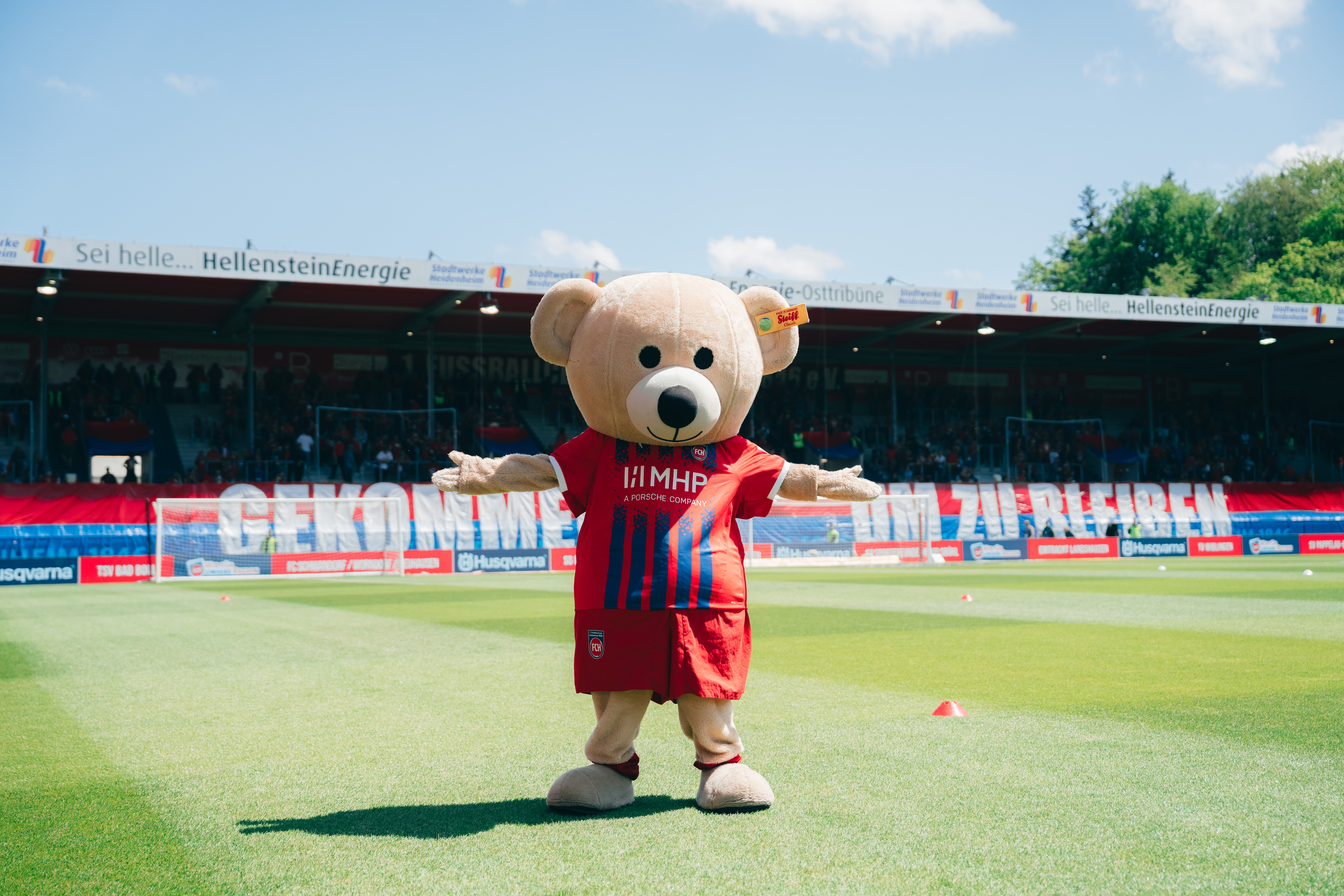 Ein Maskottchen in Form eines Bären steht auf einem Fußballfeld. Das Maskottchen trägt ein rotes Trikot mit blauen und weißen Streifen sowie eine rote Hose. Im Hintergrund sind Tribünen mit Zuschauern und Werbebanden zu sehen.