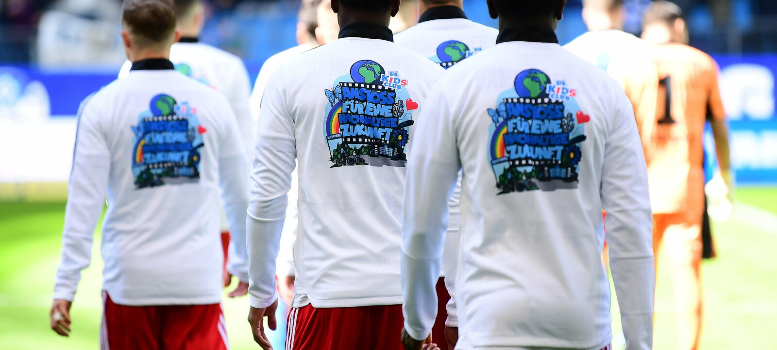 Spieler des Hamburger SV laufen mit Trikots, die das Logo "Kids Club" und eine Botschaft für Nachhaltigkeit auf dem Rücken tragen, auf den Rasen. Im Hintergrund sind Spieler des SC Paderborn 07 und Zuschauer zu sehen.