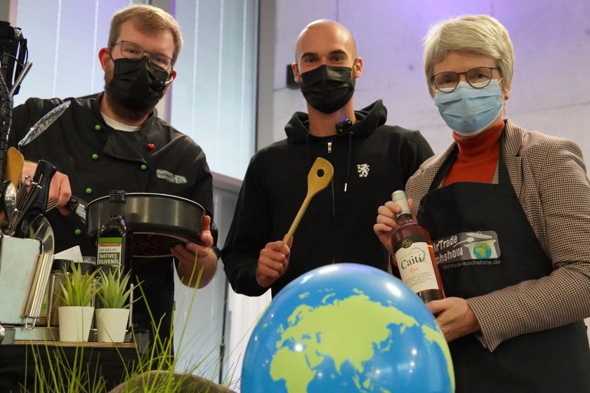 Drei Personen mit Masken, die an einer Kochshow teilnehmen. Sie halten Kochutensilien und fair gehandelte Produkte in den Händen. Im Vordergrund ist eine kleine Pflanze und ein Globus zu sehen.