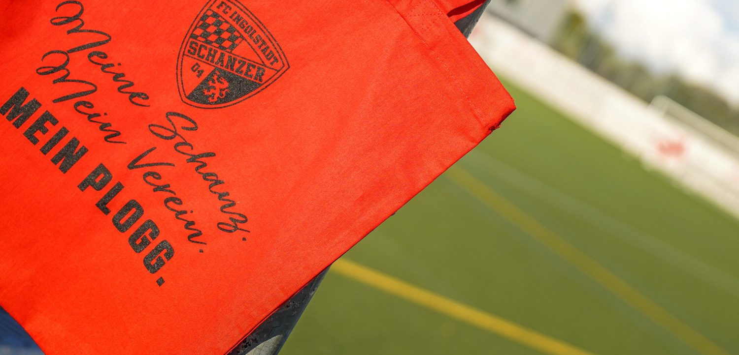 Rote Tüte mit dem Aufdruck "Meine Schanzer. Mein Verein. MEIN PLOGG." und dem Logo des FC Ingolstadt 04 auf einem Zaun vor einem Fußballfeld.