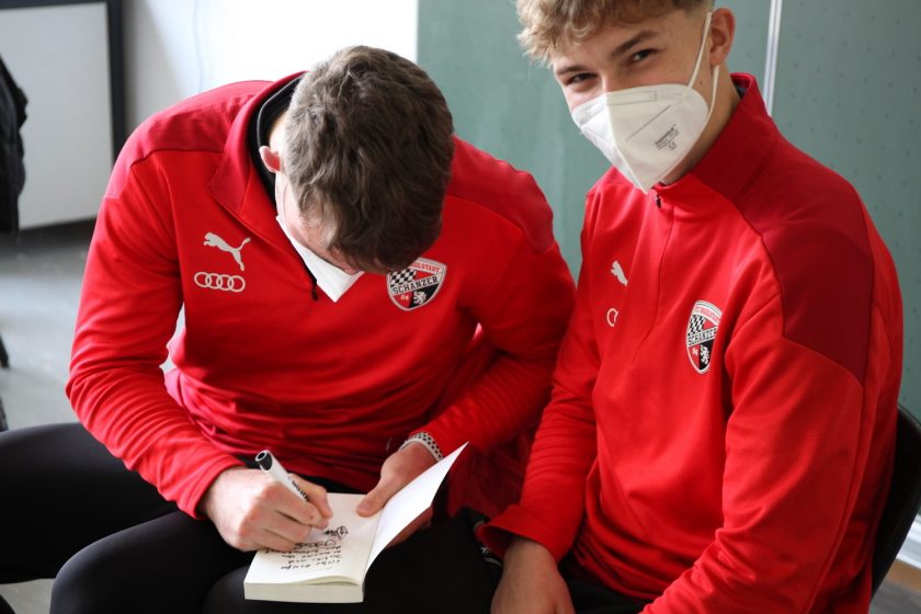 Zwei Personen in roten Jacken mit Masken sitzen nebeneinander und schreiben gemeinsam in ein Notizbuch. Beide tragen Jacken mit dem Logo des SC Freiburg und einem Audi-Logo.