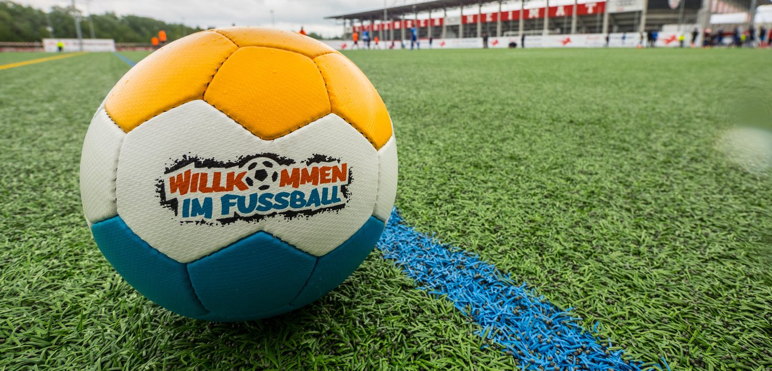 Ein orange-weißer Fußball mit dem Aufdruck "Willkommen im Fußball" liegt auf einem grünen Rasenplatz. Im Hintergrund ist eine Tribüne mit einigen Menschen zu sehen.