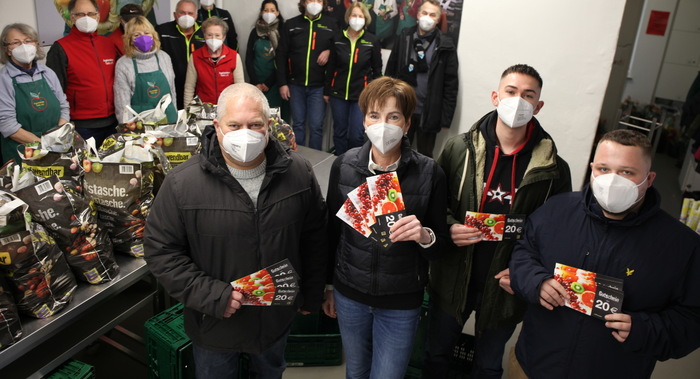 Eine Gruppe von Menschen in einem Lagerraum, die alle Masken tragen. Sie halten Samenpäckchen in den Händen und lächeln in die Kamera. Im Hintergrund sind Regale mit Säcken voller Erde und anderen Gartenzubehörteilen zu sehen.
