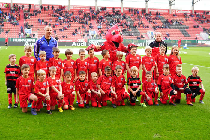 Eine Gruppe von Kindern in roten Fußballtrikots posiert auf einem Fußballfeld. Sie sind in zwei Reihen aufgestellt, mit einigen Erwachsenen und einer Mascot im Hintergrund. Die Szene findet in einem Stadion mit roten Sitzplätzen statt, und die Kinder scheinen Teil eines Jugendfußballteams oder einer ähnlichen Veranstaltung zu sein.
