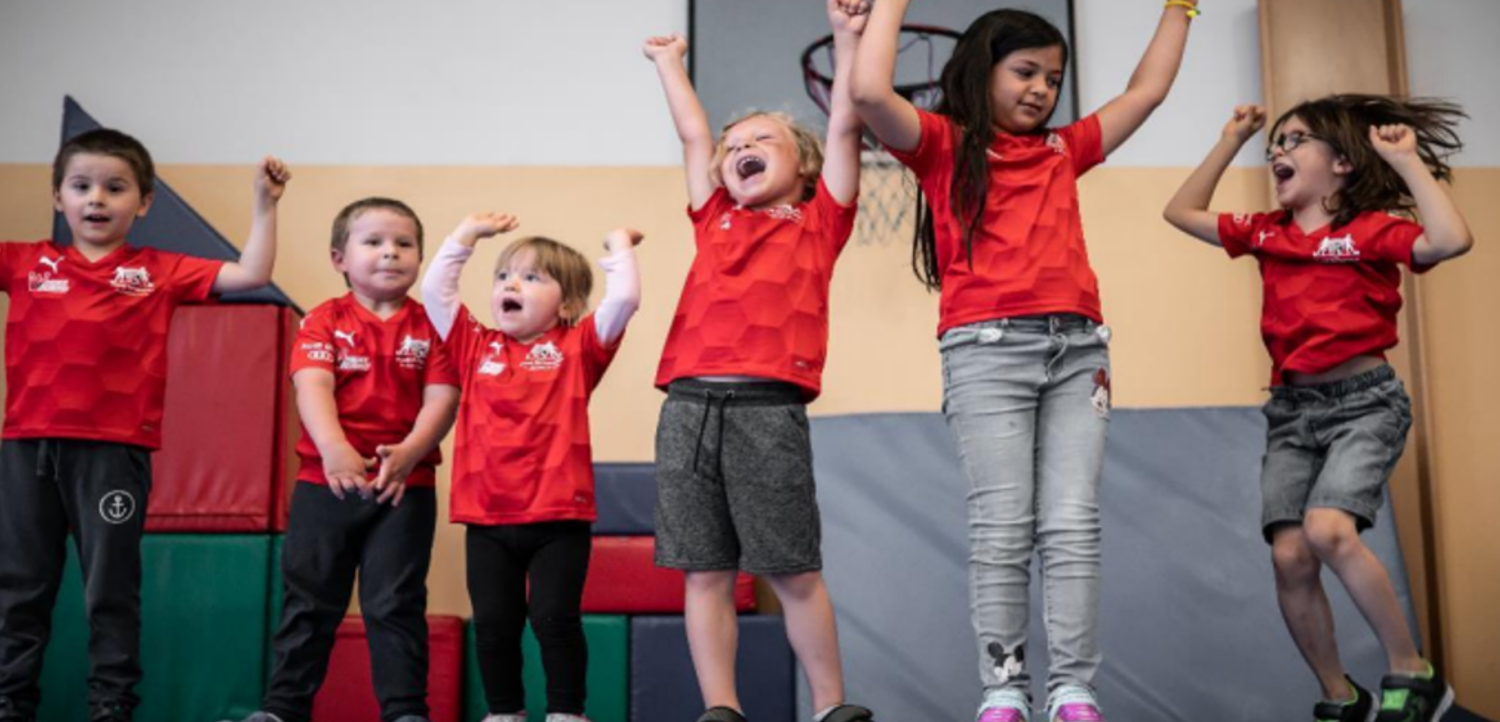 Eine Gruppe von Kindern in roten T-Shirts steht auf einer Bühne und jubelt mit erhobenen Armen. Sie scheinen sich zu freuen und sind möglicherweise Teil eines Events oder einer Feierlichkeit.