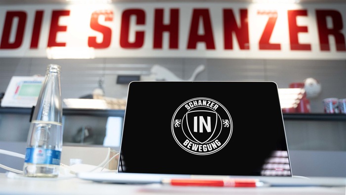 Ein offener Laptop mit dem Logo "Schanzer in Bewegung" auf dem Bildschirm. Im Hintergrund ist ein Schild mit der Aufschrift "DIE SCHANZER" zu sehen. Auf dem Tisch steht eine Flasche Wasser und ein Notizblock mit einem Stift.