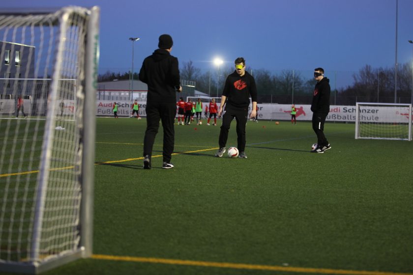 Drei Personen auf einem Fußballfeld, wobei eine Person einen Fußball kontrolliert. Die Personen tragen dunkle Kleidung und haben ihre Augen bedeckt. Im Hintergrund sind weitere Personen und ein Tor zu sehen. Die Szene findet bei Dämmerung oder in der Nacht statt, wie die künstliche Beleuchtung zeigt.