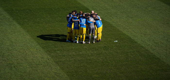Eine Gruppe von Fußballspielern in blauen und gelben Trikots steht im Kreis zusammen und feiert auf dem Rasenplatz.