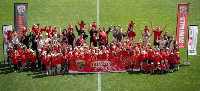 Eine große Gruppe von Kindern und Erwachsenen in roten T-Shirts und Hüten steht auf einem Fußballfeld und hält ein Banner mit dem Text "FUß MIT SCHANZI". Sie sind vor zwei Fahnen mit dem Logo "Audi Bank" und "SCHANZI" positioniert. Die Gruppe wirkt fröhlich und feiert gemeinsam.