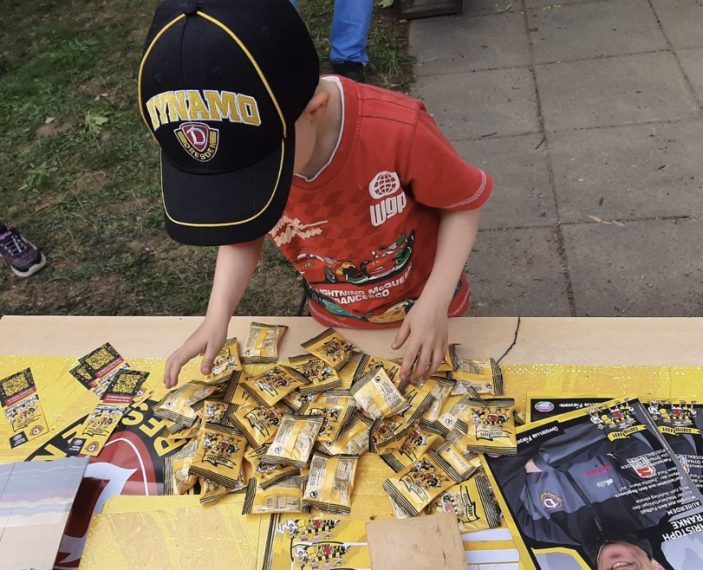Ein Kind in einem roten T-Shirt und einer schwarzen Dynamo-Kappe sortiert oder sammelt kleine gelbe Tütchen auf einem Tisch. Der Tisch ist mit Werbematerialien und einem Plakat bedeckt, auf dem ein Trikot und ein Bild eines Spielers zu sehen sind.