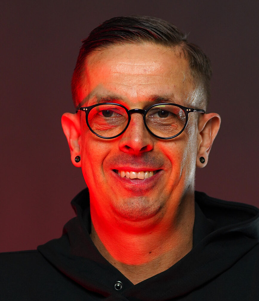 Porträtfoto eines lächelnden Mannes mit Brille und Ohrringen, der vor einem dunkelroten Hintergrund steht.
