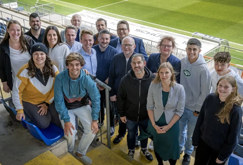 Eine Gruppe von Menschen sitzt auf den Tribünen eines Fußballstadions und lächelt in die Kamera. Die Gruppe besteht aus einer Mischung von Erwachsenen und Jugendlichen, die unterschiedlich gekleidet sind. Im Hintergrund ist der Rasen des Fußballfeldes zu sehen.