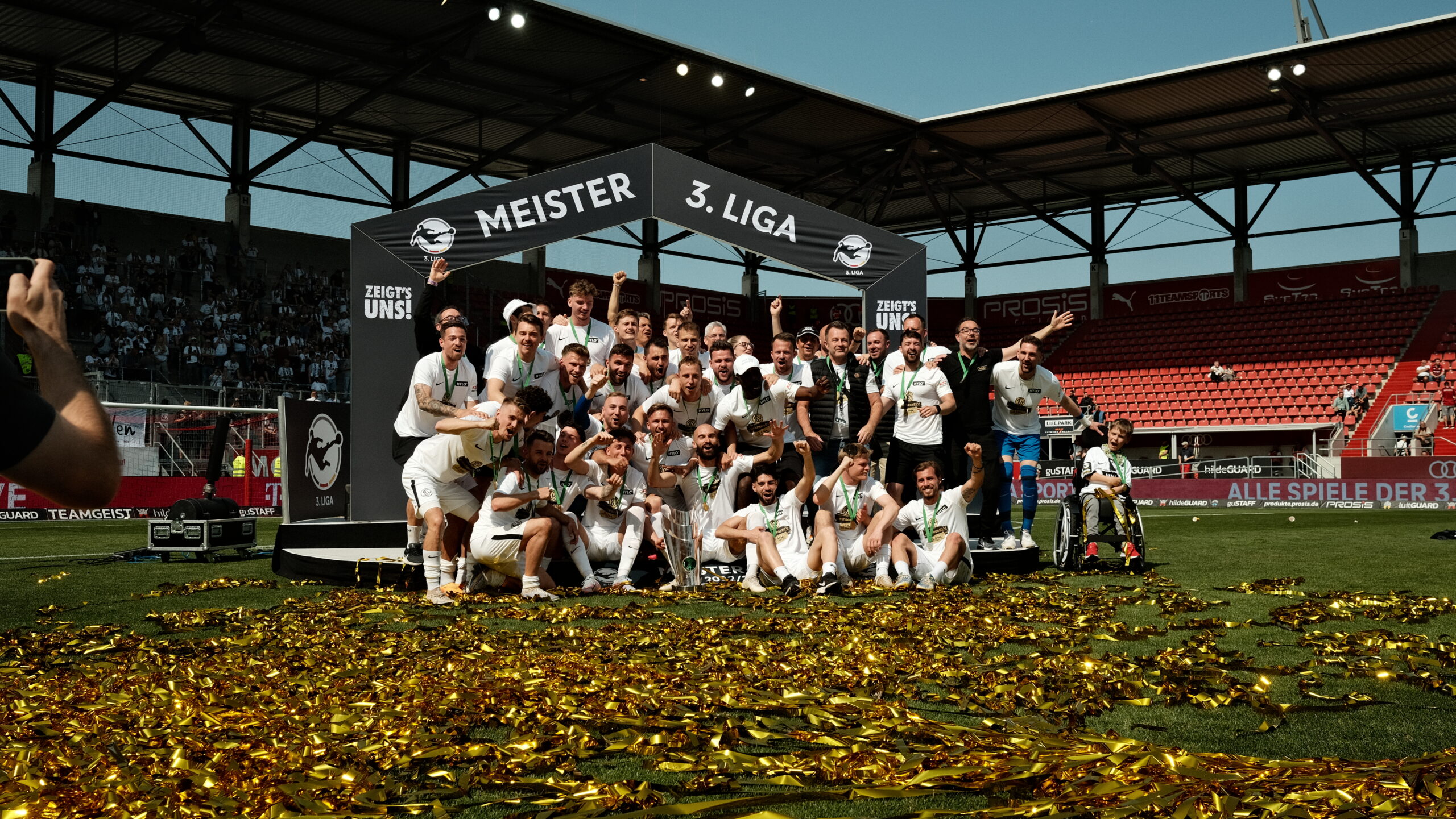 Ein Team in weißen Trikots feiert auf dem Spielfeld den Meistertitel in der 3. Liga. Die Spieler stehen und knien auf einer Bühne, die mit "Meister 3. Liga" beschriftet ist, und halten einen Pokal. Konfetti liegt auf dem Rasen, und einige Spieler halten ihre Hände hoch oder umarmen sich. Im Hintergrund sind Zuschauer auf den Tribünen zu sehen.