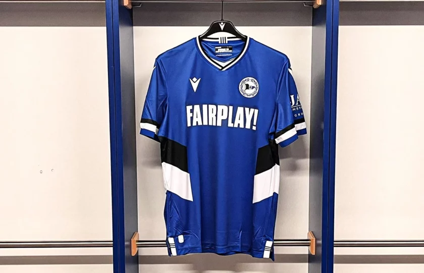 Ein blaues Fußballtrikot mit weißen und schwarzen Akzenten hängt an einem Kleiderbügel. Auf der Vorderseite des Trikots steht in großen weißen Buchstaben "FAIRPLAY!". Das Trikot hat verschiedene Logos, darunter ein rundes Logo mit einem Fußball und ein Logo mit den Buchstaben "JAG". Das Trikot ist an einem blauen Haken in einer Umkleidekabine aufgehängt.