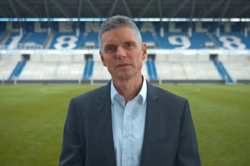 Ein Mann im Anzug steht auf einem Fußballfeld vor leeren Tribünen. Die Tribünen zeigen die Zahlen 1899, was auf den Fußballverein Karlsruher SC hinweist. Der Mann blickt direkt in die Kamera.