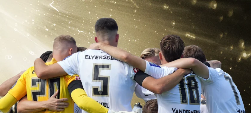 Eine Gruppe von Fußballspielern in weißen und gelben Trikots umarmen sich auf dem Spielfeld, während Konfetti und Lichter im Hintergrund zu sehen sind.