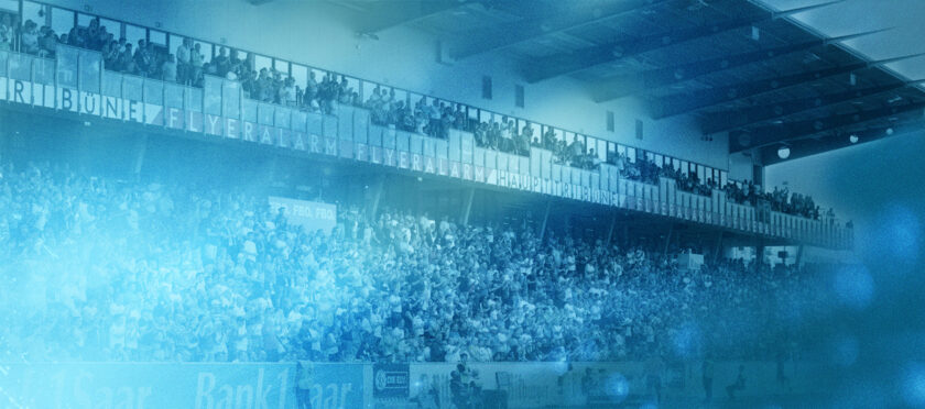 Ein Stadion voller Menschen, wahrscheinlich während eines Fußballspiels oder einer Veranstaltung. Die Haupttribüne ist mit Zuschauern gefüllt, und es gibt eine überdachte Sitzbereich-Ebene mit weiteren Zuschauern. Der Bildschirm zeigt 'FLYERALARM HAUPTTRIBÜNE' an, was auf eine Werbung oder einen Sponsor hinweist. Der Bildschirm ist leicht verschwommen, was auf eine Bewegung oder eine spezielle Effekt hinweisen könnte.