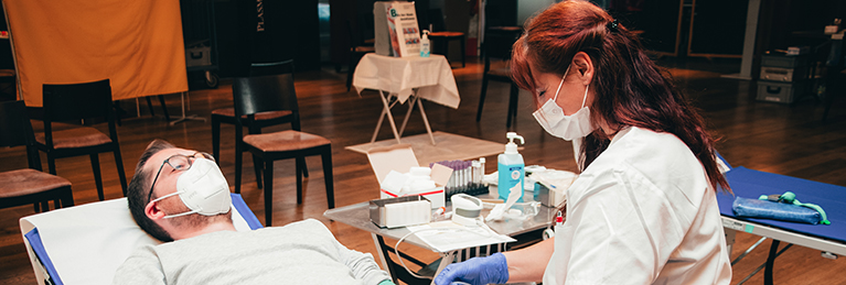 Eine medizinische Fachkraft in weißem Kittel und mit Schutzmaske nimmt einer liegenden Person, die ebenfalls eine Schutzmaske trägt, Blut ab. Im Hintergrund sind weitere Stühle und ein Tisch mit medizinischem Material zu sehen.