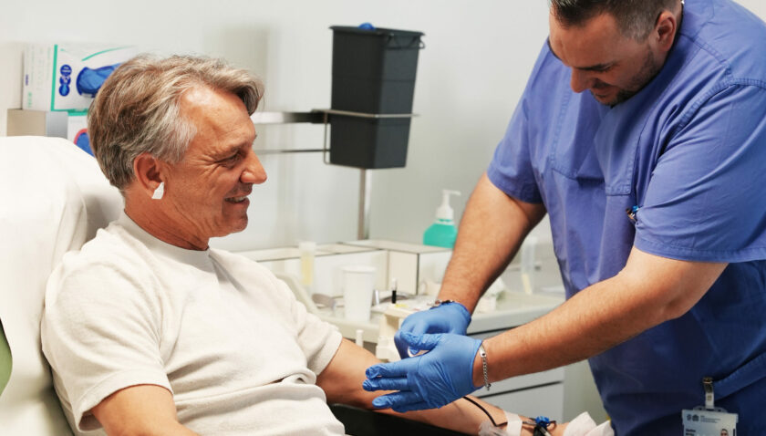 Ein älterer Mann sitzt in einem medizinischen Stuhl und lächelt, während ein medizinischer Fachmann in blauer Uniform ihm Blut abnimmt. Der Fachmann trägt Handschuhe und hält vorsichtig den Arm des Mannes fest. Im Hintergrund sind medizinische Geräte und Desinfektionsmittel zu sehen.