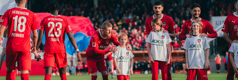 Spieler des 1. FC Heidenheim und Kinder mit ihren Betreuern auf dem Fußballfeld, alle tragen rote und weiße Trikots. Die Spieler und Kinder stehen zusammen und lächeln in die Kamera. Im Hintergrund sind Zuschauer auf den Tribünen zu sehen.