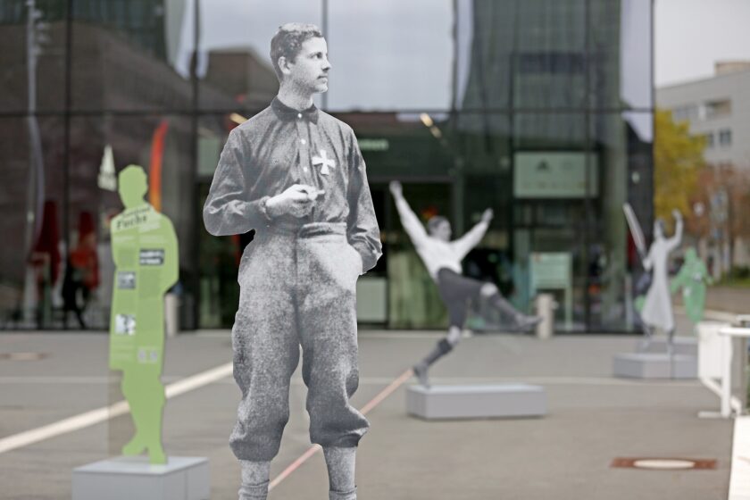 Schwarz-weiß Fotografie einer historischen Fußballfigur in traditioneller Sportkleidung, aufgestellt im öffentlichen Raum vor einem modernen Gebäude. Im Hintergrund sind weitere Figuren und Passanten zu sehen, die verschiedene Posen einnehmen.