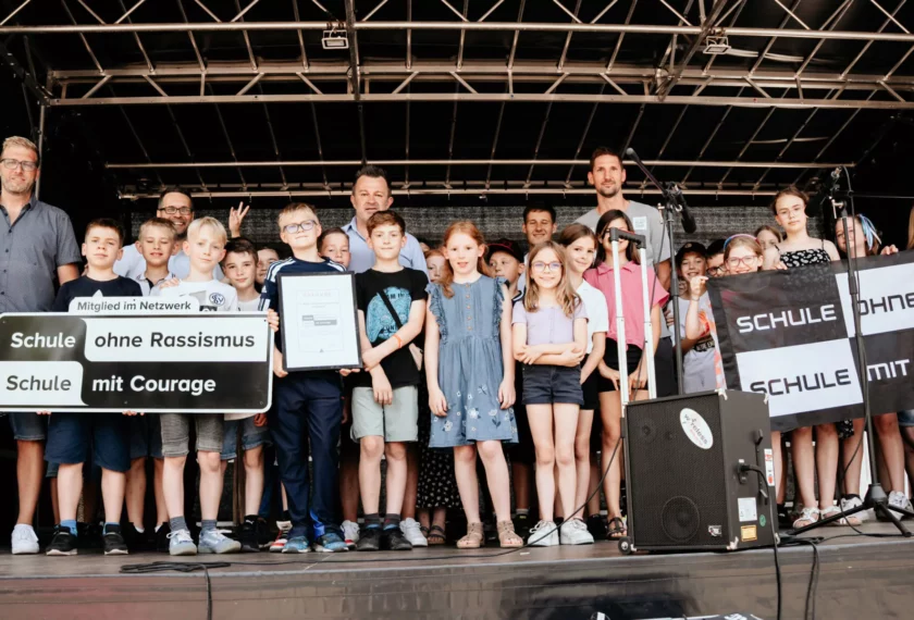 Eine Gruppe von Kindern und Erwachsenen steht auf einer Bühne und hält Schilder mit der Aufschrift "Schule ohne Rassismus - Schule mit Courage". Ein Kind in der Mitte hält eine Urkunde oder ein Zertifikat. Die Veranstaltung scheint eine Feier oder eine Auszeichnung für das Engagement gegen Rassismus und für Courage zu sein.