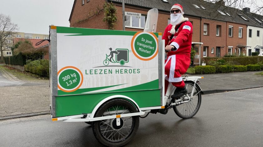 Ein Mann in Weihnachtsmann-Kostüm fährt ein Lastenfahrrad mit der Aufschrift "LEEZEN HEROES" und "So jovel transportiert Münster!". Das Fahrrad hat eine Tragfähigkeit von 300 kg und ein Ladevolumen von 1,3 m³. Im Hintergrund sind Wohnhäuser zu sehen.