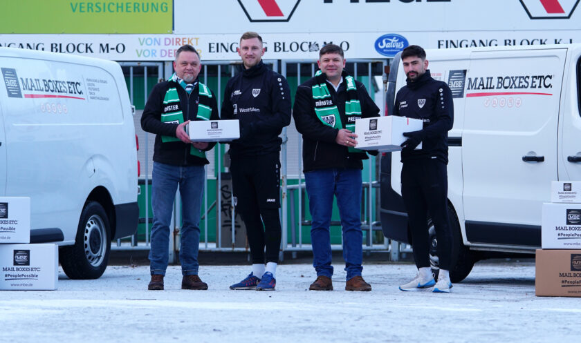 Vier Personen stehen vor einem weißen Lieferwagen mit der Aufschrift "MAIL BOXES ETC." Sie halten jeweils eine Schachtel in den Händen und tragen schwarze Jacken mit grünen und weißen Schals. Der Hintergrund zeigt ein Stadion mit Schildern und Werbung.