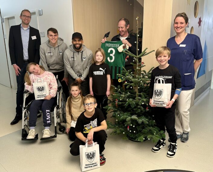 Eine Gruppe von Menschen, darunter Fußballspieler und Kinder, posieren für ein Foto auf einer Kinderstation. Die Kinder halten Tüten mit dem Logo des SC Preußen Münster, während ein Spieler ein grünes Trikot mit der Nummer 3 hochhält. Im Hintergrund steht ein geschmückter Weihnachtsbaum.