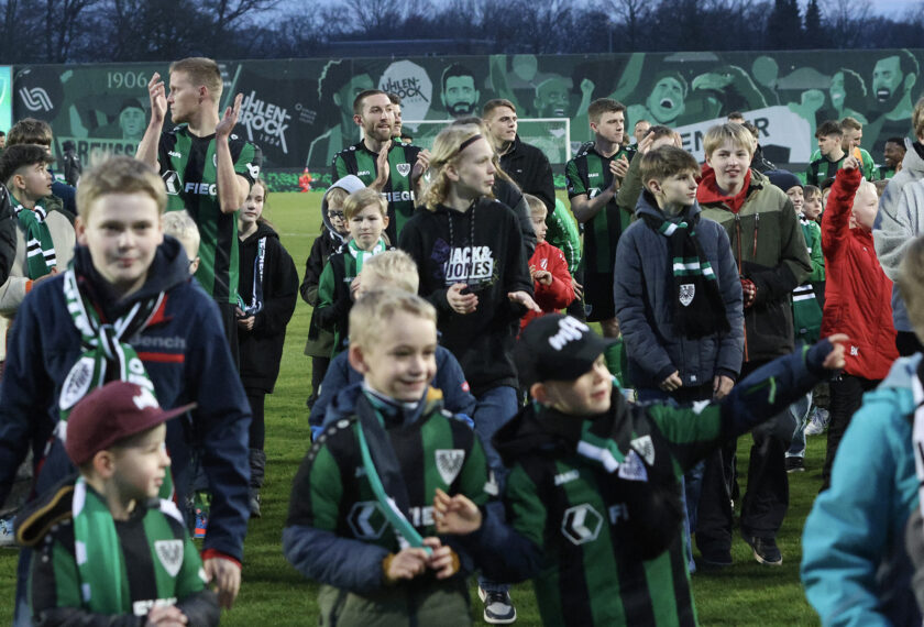 Eine Gruppe von Kindern und Erwachsenen, darunter Fußballspieler in grün-schwarzen Trikots, stehen auf einem Fußballfeld. Die Kinder tragen Schals und Mützen mit dem Vereinslogo und sind sichtlich begeistert. Im Hintergrund ist eine Wand mit Graffiti und dem Schriftzug "Uhlenhorsts" zu sehen. Die Szene wirkt fröhlich und gesellig, möglicherweise handelt es sich um ein Event oder eine Feierlichkeit im Zusammenhang mit dem Fußballverein.