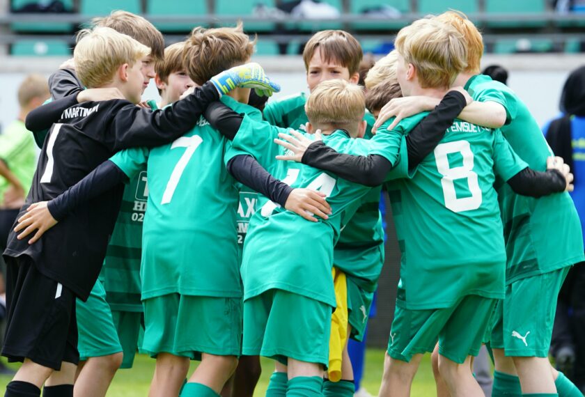 Eine Gruppe von jungen Fußballspielern in grünen Trikots umarmen sich auf dem Spielfeld, feiernd und sich gegenseitig unterstützend. Die Spieler tragen die Nummern 1, 7, 8 und 21 auf ihren Trikots. Sie scheinen sich über einen Erfolg oder ein Tor zu freuen.